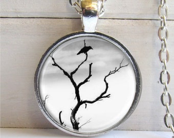 May include: A silver necklace with a round pendant featuring a black and white image of a crow perched on a bare tree branch against a cloudy sky.