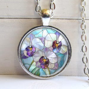 May include: A silver-toned pendant necklace featuring a round, stained-glass style design. The pendant showcases three orchid flowers in shades of purple, white, and yellow against a light blue background. The chain is also silver-toned.