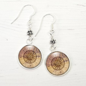 May include: A pair of silver dangle earrings with a compass design. The compass is on a brown background with a vintage look. The earrings have a silver bead at the top.