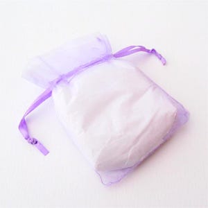 May include: A small, sheer lavender gift bag filled with white contents. The bag is made of a delicate, translucent fabric and is tied with a matching lavender ribbon. The bag is on a white background.