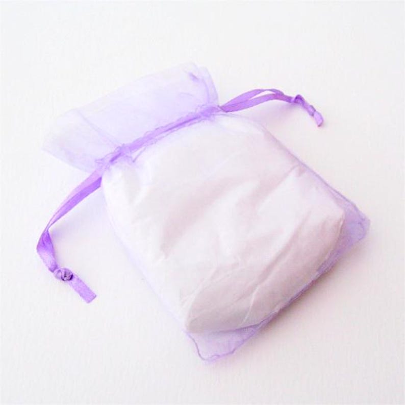 May include: A small, sheer lavender gift bag filled with white contents. The bag is made of a delicate, translucent fabric and is tied with a matching lavender ribbon. The bag is on a white background.