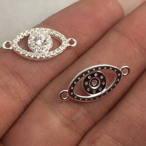 Pode incluir: Dois pingentes de olho de prata. Um pingente tem um strass transparente no centro rodeado por strass menores. O outro pingente tem um strass transparente no centro com um círculo prateado ao redor.