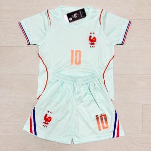 May include: A light mint green short-sleeved jersey and matching shorts set featuring the number 10 in orange, a red rooster emblem with two stars above it, and blue, white, and red striped trim on the sleeves and shorts. A black tag reads FRANCE.