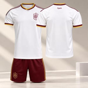 Può includere: Una maglia da calcio bianca a maniche corte con colletto e bordi delle maniche bordeaux e oro. Il davanti presenta uno stemma, mentre il retro riporta la scritta España. Il set include pantaloncini bordeaux con strisce laterali dorate.