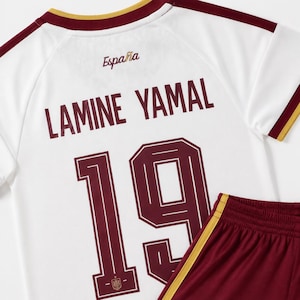 May include: A white soccer jersey with maroon and gold trim, featuring the text Espa&ntilde;a, the name Lamine Yamal, and the number 19 in maroon. The jersey is paired with maroon soccer shorts that have a gold stripe along the side seam.