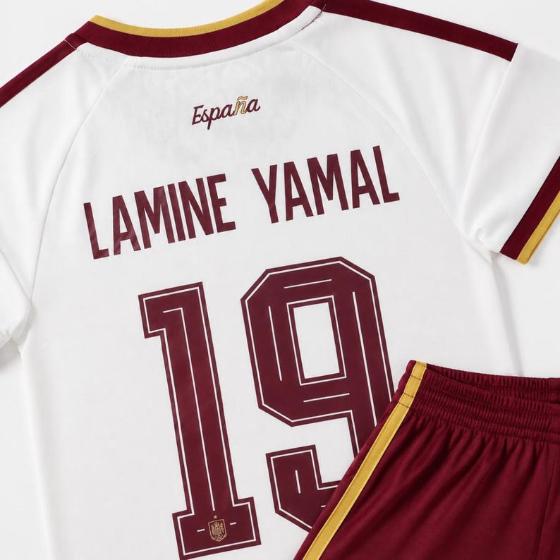 May include: A white soccer jersey with maroon and gold trim, featuring the text Espa&ntilde;a, the name Lamine Yamal, and the number 19 in maroon. The jersey is paired with maroon soccer shorts that have a gold stripe along the side seam.