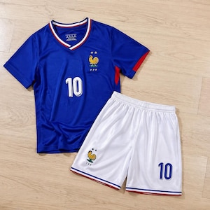 Può includere: Una maglia da calcio blu con scollo a V e bordini bianchi e rossi, con il numero 10 in bianco, un emblema di gallo giallo e due stelle. Abbinata a pantaloncini da calcio bianchi con bordini rossi e blu, il numero 10 e un emblema di gallo.