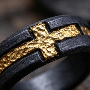 May include: A ring featuring a dark, matte black band with a textured surface. An inlaid gold-colored cross with a rough, hammered finish runs horizontally along the center of the band, creating a distinct contrast between the dark and metallic elements.