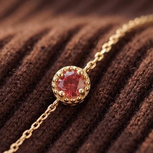 Op de afbeelding: Een delicate gouden kettingarmband met een enkele ronde, gefacetteerde rode edelsteen in een ronde gouden zetting met een kralenrand. De zetting is direct in de fijne schakelketting geïntegreerd, wat zorgt voor een verfijnd sieraad.