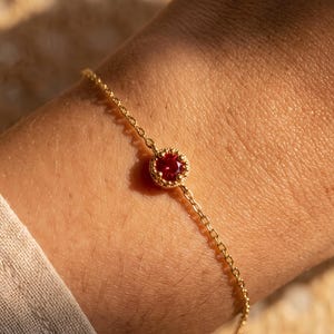 May include: A delicate gold chain bracelet featuring a single, round, faceted red gemstone set in a textured, beaded gold bezel setting. The bracelet is secured around the wrist, showcasing the vibrant red stone and the fine, polished gold links.