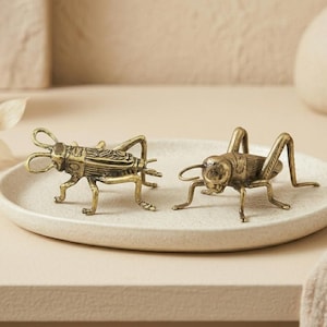 May include: Two brass-coloured metal cricket figurines with detailed textured bodies, long antennae, and bent legs, positioned side-by-side on a round, off-white ceramic plate with a speckled finish and a slightly raised edge.