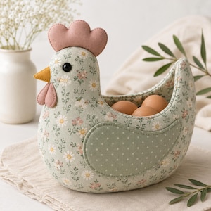 May include: A fabric chicken-shaped basket featuring a light green base with a floral pattern of white and pink flowers. The chicken has a solid pink comb and wattle, a yellow beak, and a solid green wing with white polka dots, holding three brown eggs.
