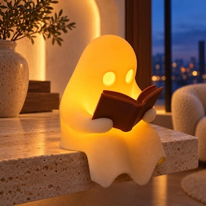 May include: A glowing, off-white ghost-shaped lamp sitting on an edge, holding a small, dark brown open book. The lamp emits a warm, soft yellow light from within, illuminating its rounded form and the book it holds in its hands.