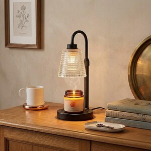 May include: A black metal candle warmer lamp with a clear, textured glass shade. The lamp sits on a circular base, heating a small amber glass jar candle with a visible flame and wisps of smoke rising from the wick into the lamp shade.