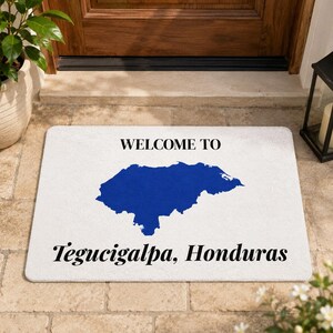 May include: A rectangular white doormat featuring the text "WELCOME TO" in black, a solid blue silhouette map of Honduras in the center, and the text "Tegucigalpa, Honduras" in black at the bottom, designed for placement at an entryway.
