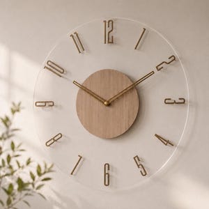Puede incluir: Un reloj de pared redondo con una esfera de acrílico transparente, números metálicos en tono dorado y manecillas a juego. Un panel circular de chapa de madera clara está centrado en la esfera, creando un contraste con el fondo de acrílico.