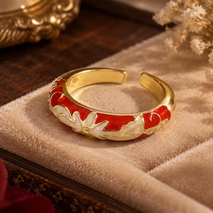 Puede incluir: Un anillo abierto de tono dorado con un vibrante patrón floral de esmalte rojo y blanco. La banda tiene un acabado liso y pulido con un diseño continuo de pétalos y hojas estilizadas que envuelven el exterior de la banda de metal.