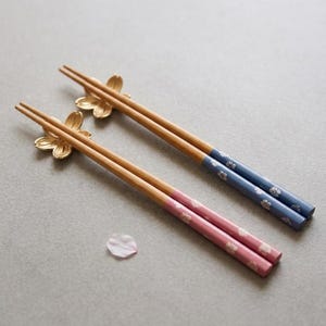 以下が含まれることがあります： 金色の花形の金属製箸置きが付いた木製の箸2膳。一膳は青い持ち手に白い花柄が施され、もう一膳はピンクの持ち手に白い花柄が施されています。近くには小さな白い花びらが一枚置かれています。