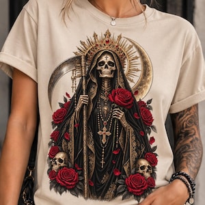 Puede incluir: Una camiseta beige con un estampado gráfico de una figura esquelética con túnica negra con capucha, corona y collares de cruz superpuestos. La figura sostiene una guadaña y una rosa roja, rodeada de rosas rojas adicionales y dos calaveras.