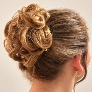 Puede incluir: Un postizo de moño desordenado en una paleta de colores castaño claro y rubio con reflejos, peinado en una forma circular voluminosa y texturizada. El postizo está sujeto al cabello natural recogido hacia atrás y luce un pequeño pendiente dorado.