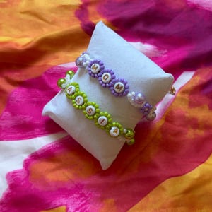Peut inclure: Deux bracelets en perles avec des perles en forme de fleur et des perles lettres rondes. Le bracelet du haut est violet avec le texte MOM, et le bracelet du bas est vert citron avec le texte ASSIA. Ils incluent de petites perles blanches.