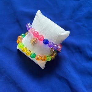 Peut inclure: Deux bracelets extensibles composés de perles sphériques translucides aux couleurs de l'arc-en-ciel. Chaque bracelet comporte une petite breloque en forme de tortue dorée suspendue au rang de perles, posé sur un coussin de présentation blanc.