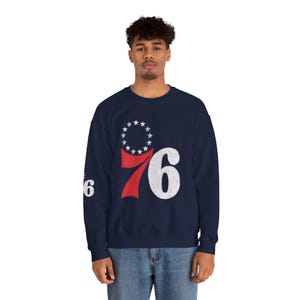 May include: A navy blue crewneck sweatshirt featuring a large graphic on the chest with the number 76 in red and white, topped by a circle of thirteen white stars. A smaller version of the 76 graphic is printed on the right sleeve of the garment.