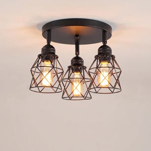 May include: A matte black ceiling light fixture featuring a circular base plate and three adjustable pendant lights. Each light has a decorative metal wire cage shade surrounding a glowing Edison-style bulb, creating a focused illumination pattern.