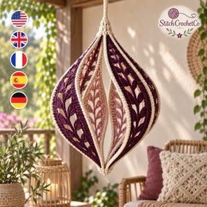 Op de afbeelding: Een hangend gehaakt ornament in de vorm van een druppel met afwisselende panelen van donkerpaars en lichtroze garen. Het ontwerp bevat ingewikkelde bladpatronen omlijnd met crèmekleurig garen. De tekst StitchCrochetCo is zichtbaar.