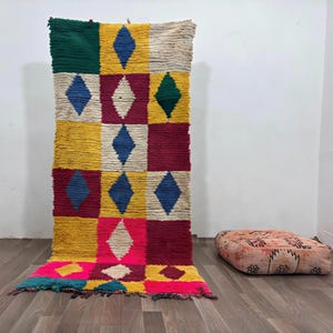 Peut inclure: Un tapis rectangulaire en laine composé d'une grille de blocs colorés en jaune, rouge foncé, vert, crème et rose vif, chacun contenant un losange centré d'une couleur contrastante. Un coussin de sol carré à motifs est posé à proximité.