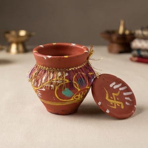 May include: A terracotta pot with a matching lid, featuring hand-painted yellow, white, and teal floral patterns. The rim is decorated with a gold and purple fringe trim. The lid displays a hand-painted yellow swastika symbol and white accents.