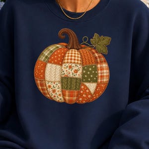 May include: A navy blue long-sleeve sweatshirt featuring a large central graphic of a pumpkin composed of various patchwork patterns in shades of orange, green, and white, including plaid, floral, and cable knit textures with a brown stem.