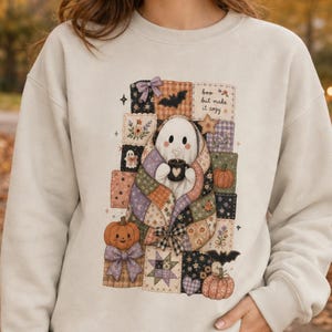 Op de afbeelding: Een beige sweater met ronde hals en een afbeelding van een geest gewikkeld in een patchwork deken, die een mok vasthoudt. Het ontwerp bevat pompoenen, vleermuizen, sterren en de tekst 'boo but make it cozy' omringd door patronen.