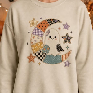 Puede incluir: Una sudadera de cuello redondo en color beige con un gráfico central de una luna creciente y un fantasma. La luna está compuesta por varios patrones de patchwork que incluyen lunares, cuadros vichy, flores y cuadros en tonos naranja, morado y azul.