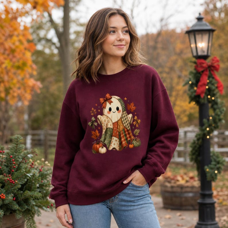 Op de afbeelding: Een bordeauxrood sweatshirt met ronde hals en een centrale afbeelding van een geest gemaakt van patchwork-stofpatronen. De geest is versierd met een klein strikje en omringd door herfstbladeren, kleine pompoenen en strikjes in oranje en bruine tinten.