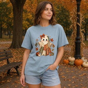 Op de afbeelding: Een lichtblauw t-shirt met korte mouwen en een centrale afbeelding van een geest gemaakt van patchwork-stofpatronen, versierd met een strik, omringd door herfstbladeren, kleine strikjes en pompoenen in oranje, groene en bruine tinten.