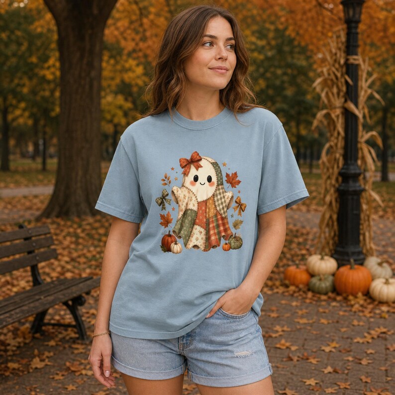 Op de afbeelding: Een lichtblauw t-shirt met korte mouwen en een centrale afbeelding van een geest gemaakt van patchwork-stofpatronen, versierd met een strik, omringd door herfstbladeren, kleine strikjes en pompoenen in oranje, groene en bruine tinten.