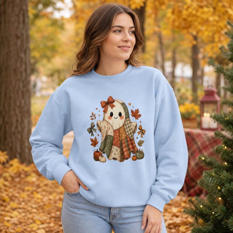 Op de afbeelding: Een lichtblauw sweatshirt met ronde hals en een centrale afbeelding van een geest gemaakt van verschillende patchwork-stofpatronen. De geest is versierd met een klein strikje en omringd door herfstbladeren, kleine pompoenen en decoratieve strikken.