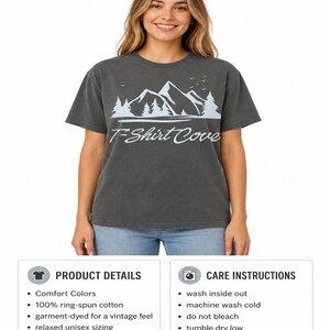 Op de afbeelding: Een antracietgrijs T-shirt met korte mouwen en een lichtblauwe afbeelding van bergtoppen, dennenbomen en vogels boven de tekst T-Shirt Cove in een cursief lettertype. Het shirt heeft een ronde hals en een relaxte pasvorm.