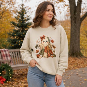 Op de afbeelding: Een beige sweater met ronde hals en een centrale afbeelding van een geest gemaakt van patchwork-stofpatronen. De geest draagt een rode strik en is omringd door herfstbladeren, kleine pompoenen en decoratieve strikken in oranje en groene tinten.