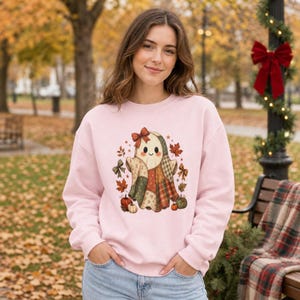 Op de afbeelding: Een lichtroze sweatshirt met lange mouwen en een centrale afbeelding van een geest die een deken in patchworkstijl draagt. De geest is omringd door herfstbladeren, kleine pompoenen en decoratieve strikken in tinten oranje, bruin en groen.