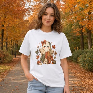 Op de afbeelding: Een wit T-shirt met korte mouwen en een centrale afbeelding van een geest gemaakt van patchworkstof in herfstkleuren. De geest heeft een strik op het hoofd en is omringd door kleine pompoenen, esdoornbladeren en decoratieve strikken in oranje, bruine en groene tinten.