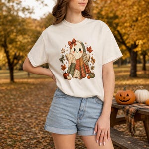 Op de afbeelding: Een cr&egrave;mekleurig T-shirt met korte mouwen en een centrale afbeelding van een lachende geest met een rode strik, versierd met een patchworkpatroon in herfstkleuren, omringd door kleine pompoenen en vallende esdoornbladeren in oranje en bruine tinten.