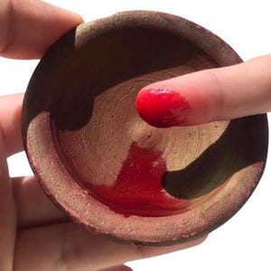 May include: A small, round, brown terracotta bowl with a textured, matte surface. The interior of the bowl contains a pool of vibrant, glossy red liquid, with a finger coated in the same red substance resting inside the bowl's basin.