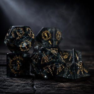 May include: A set of polyhedral dice made from dark, translucent, marbled resin with gold-colored engraved markings. The dice feature intricate, swirling patterns within the material and sharp, clear numbers etched into each face of the various shapes.