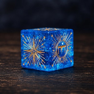 Puede incluir: Un dado cúbico azul translúcido con intrincados grabados de color dorado. Una cara muestra un gran diseño de estrella de múltiples puntas, mientras que la cara adyacente presenta un emblema circular con una letra hebrea central rodeada de líneas radiantes.