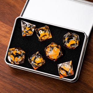 May include: A set of seven polyhedral dice made of clear resin with embedded orange inclusions and white numbering. The dice are arranged in a rectangular metal storage tin with a black foam insert, featuring various shapes including d20, d12, d10, d8, d6, and d4.