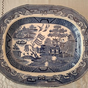 Pode incluir: Uma travessa oval de cerâmica com um padrão de salgueiro em azul e branco. O design retrata uma paisagem com um pagode, uma ponte, salgueiros e dois pássaros em voo. A borda larga é decorada com intrincados motivos geométricos e florais.