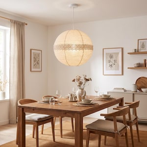 May include: A spherical paper pendant light fixture featuring a horizontal band of intricate cream-colored lace trim around the center. The lamp is suspended from a white ceiling mount by a thin white cord, emitting a soft, warm glow.