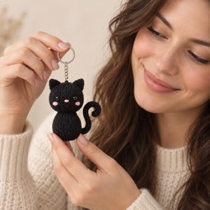 Può includere: Un portachiavi a forma di gatto lavorato a maglia nero con testa e corpo rotondi, due orecchie a punta, una coda arricciata, piccoli occhi neri, guance ricamate in rosa e un naso rosa. Il gatto è fissato a un anello in metallo argentato con catena.